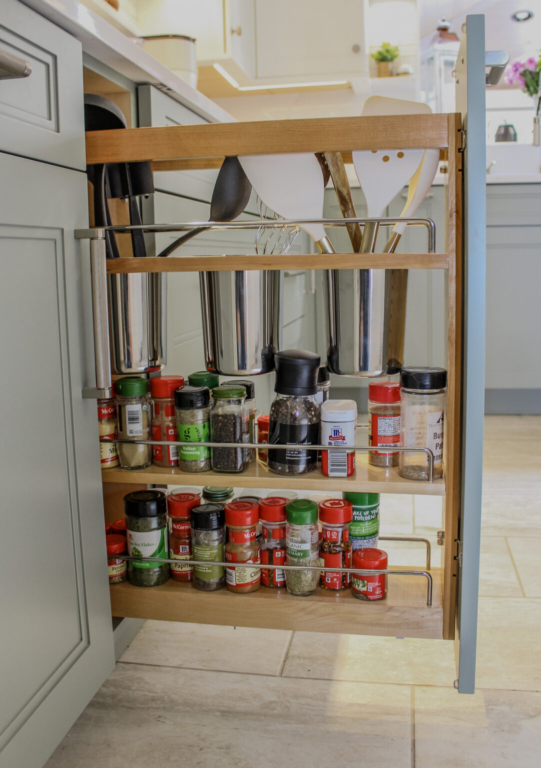 Base pullout utensil organizer in kitchen layout. Cabinet made of maple painted in the acacia haze finish with Austin door style, full of kitchen utensils and spices.