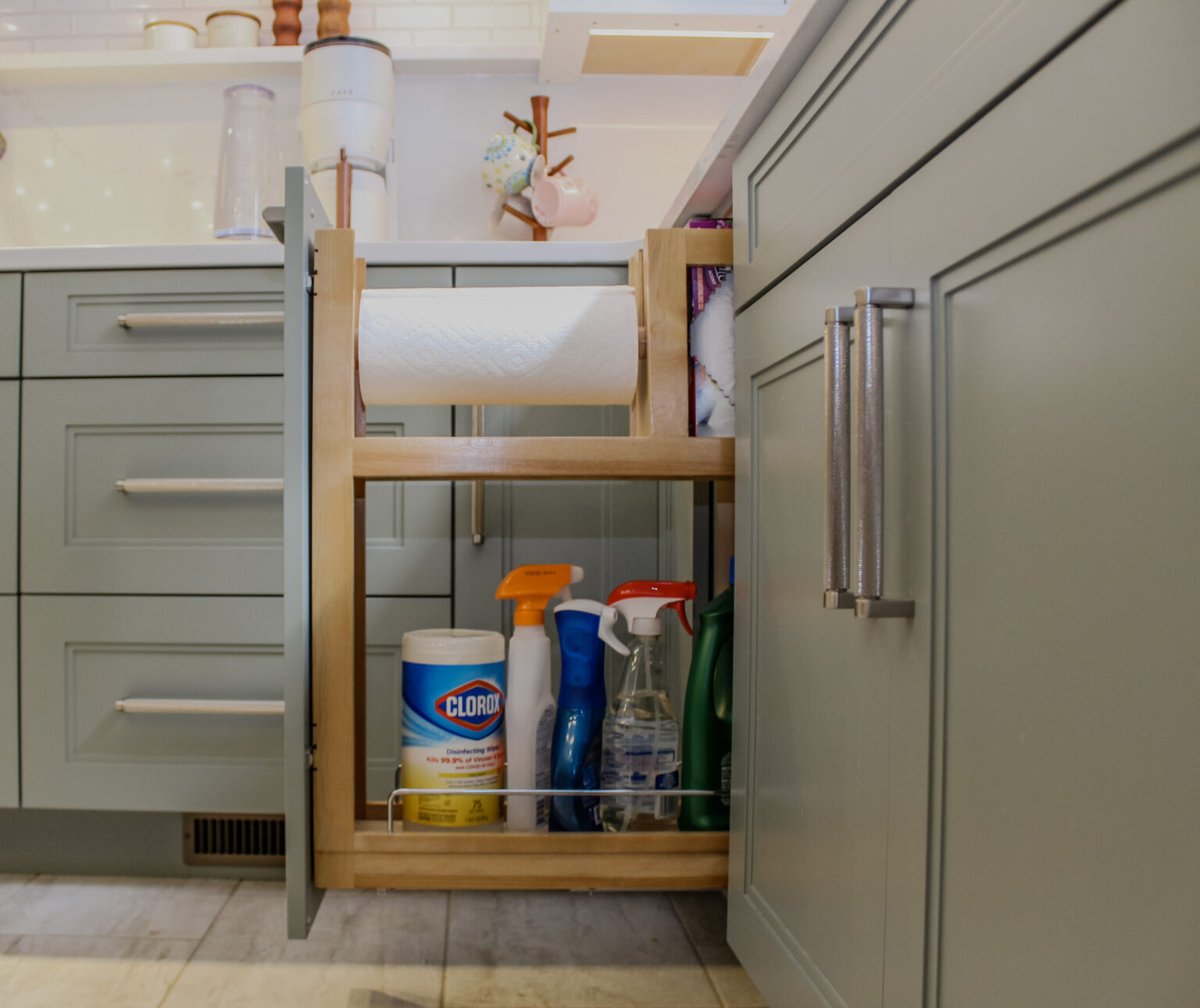 Paper towel storage pullout cabinet. Cabinet is made of maple painted in the acacia haze finish and features Austin door style, filled with paper towel on dowel and cleaning supplies underneath.